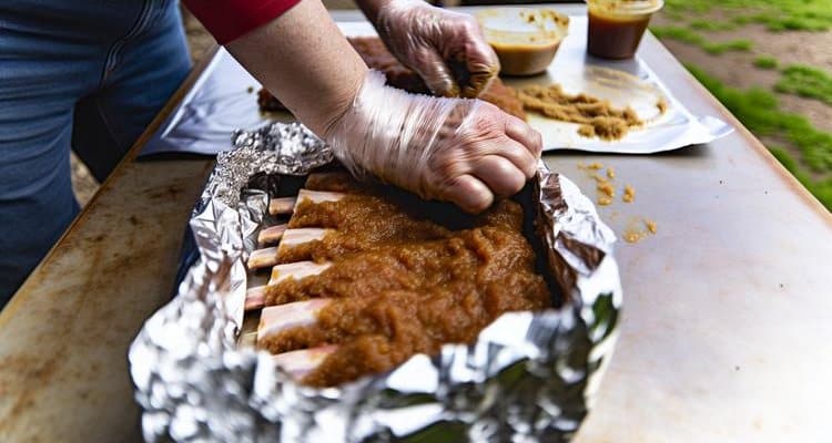 Hands wrapping smoked pork ribs in aluminum foil with visible seasonings for Phase 2 of 3-2-1 method