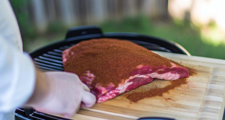 applying brisket rub to raw beef brisket on cutting board, generous coating of reddish-brown spice rub visible - cooking process