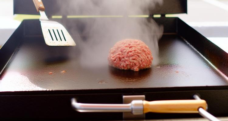 Ground beef ball on Blackstone griddle surface before smashing with spatula nearby