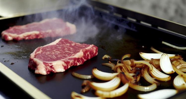 Ribeye cooking on flat-top griddle with metal spatulas