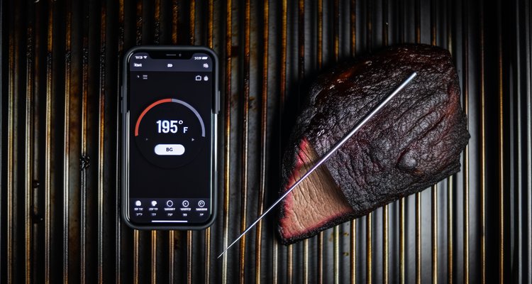 Wireless meat thermometer probe inserted in brisket on smoker showing 195°F on smartphone app
