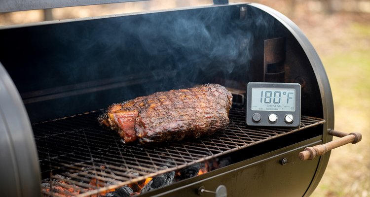 Smoked brisket on a smoker grate with a digital thermometer reading stuck in the stall zone, surrounded by smoke in a rustic outdoor BBQ setting
