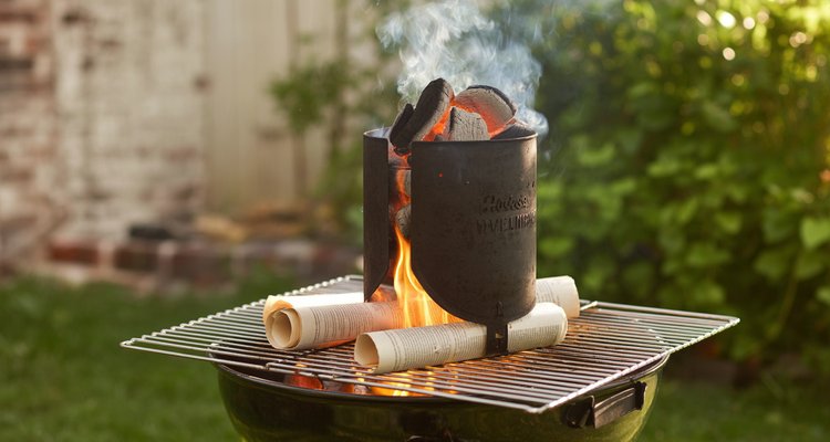Charcoal chimney starter sitting on grill grates with newspaper burning underneath and smoke rising