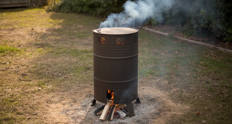 55-gallon steel drum being used as a charcoal kiln with smoke rising from vent holes and fire at the base in a backyard setting