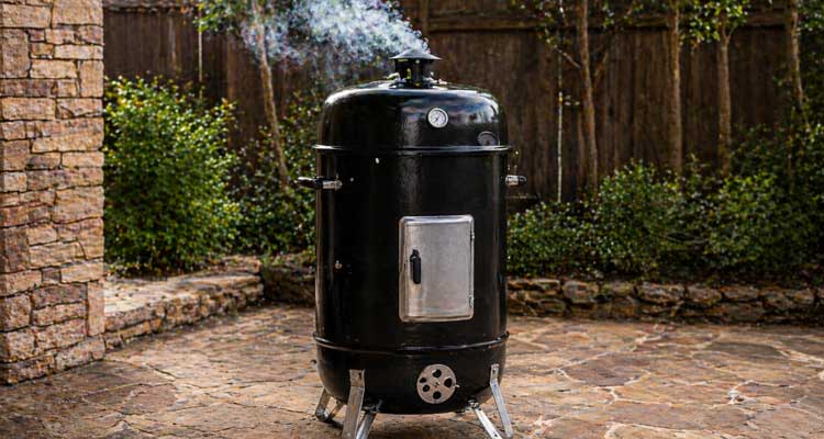 Black cylindrical charcoal smoker on patio with smoke trailing from the top vent