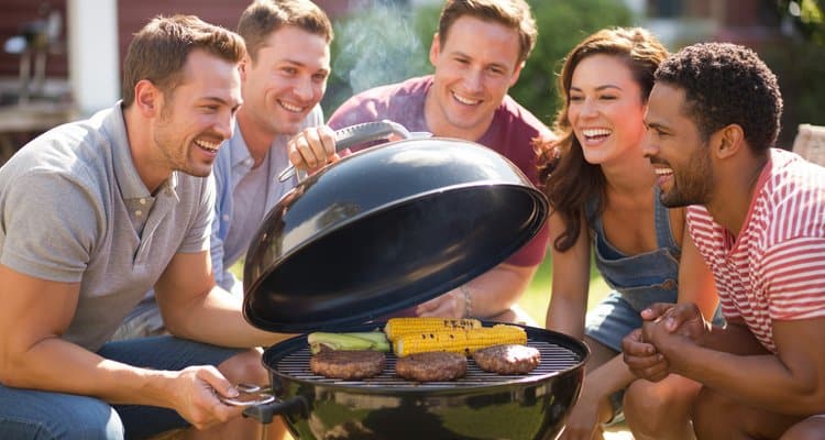 Friends gathered around a charcoal barbeque grill on a sunny backyard patio lifting the lid to reveal perfectly grilled food