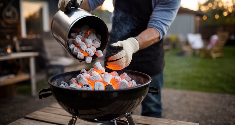 Person wearing heat-resistant gloves pouring glowing hot charcoal from a chimney starter into a kettle grill in a backyard setting