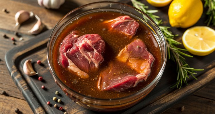 Steak marinating in glass bowl with herbs and garlic Raw steak pieces marinating in a glass bowl with soy sauce, garlic cloves, fresh rosemary, lemon, and peppercorns on a dark wooden cutting board