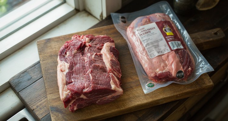 Raw beef brisket and vacuum-sealed corned beef brisket side by side on a wooden cutting board showing the color difference between uncured and cured meat