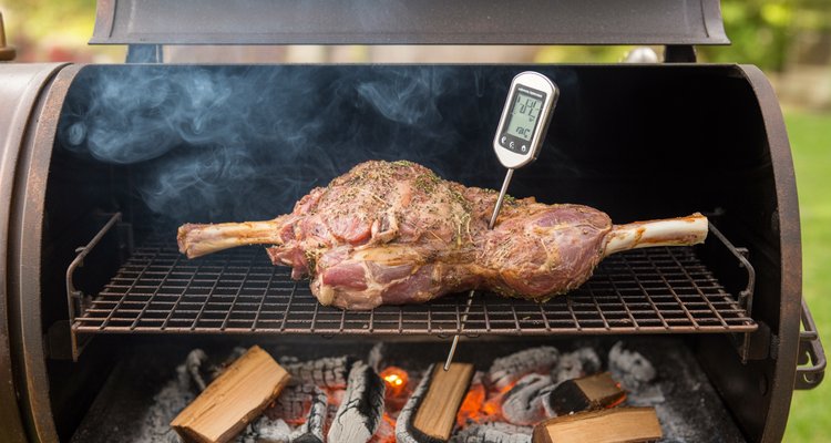 Seasoned leg of lamb smoking inside a smoker with thermometer probe and wood chunks