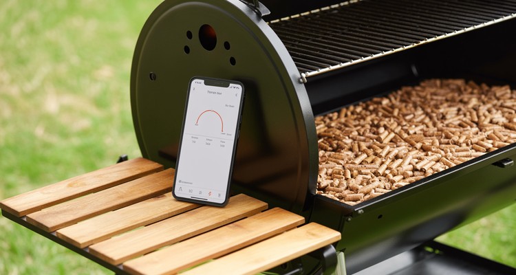 Smartphone showing digital temperature graph app resting next to a grill hopper filled with natural hardwood BBQ pellets outdoor lighting