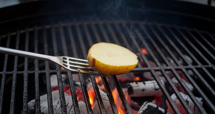 Halved raw potato on a grilling fork being rubbed across hot cast iron grill grates over glowing charcoal
