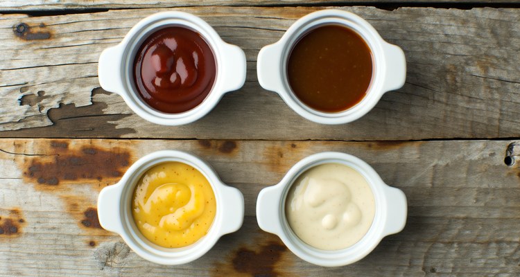 Four ramekins of different regional BBQ sauces — vinegar, tomato, mustard, and white — arranged on a rustic wood surface