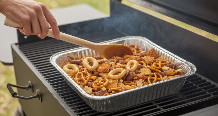 Hand stirring smoked Chex Mix with a wooden spoon in an aluminum pan on a smoker