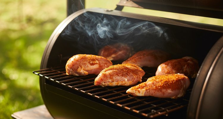 Seasoned raw chicken breasts with spice rub on smoker grates with white smoke rising