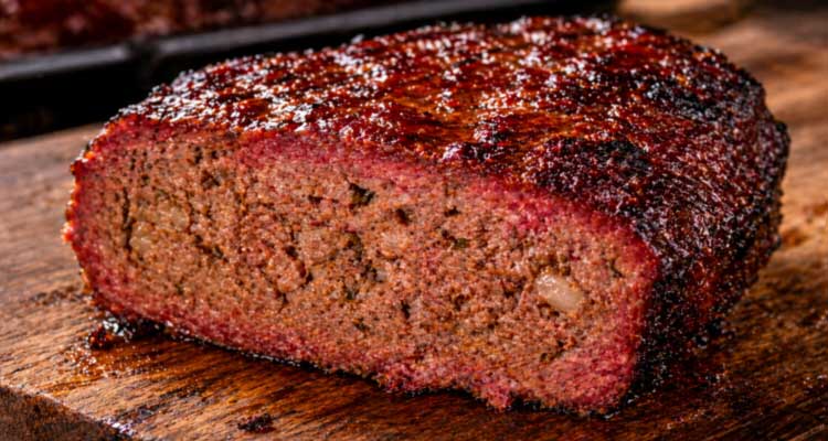 Thick slice of smoked meatloaf with a caramelized BBQ glaze and pink smoke ring on a rustic wooden cutting board