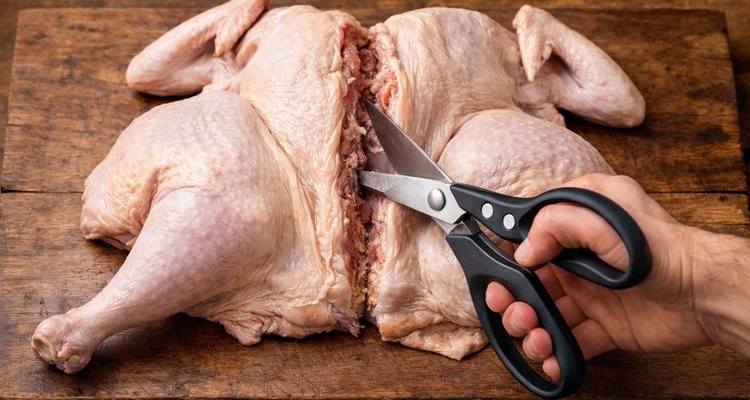 Two hands holding kitchen scissors positioned along the spine of a whole uncooked turkey that is lying breast-side down on a wooden cutting board.