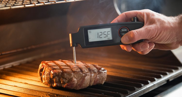 Hand inserting instant-read meat thermometer horizontally into a steak on a grill showing 125°F pull-off temperature