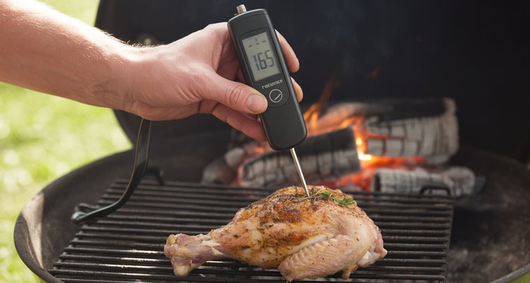 Hand inserting a digital instant-read thermometer into a bone-in chicken thigh on a charcoal grill showing 165°F safe internal temperature