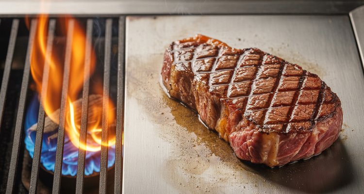 Perfectly seared ribeye steak with grill marks resting on cool indirect zone beside visible flames on direct heat side of gas grill