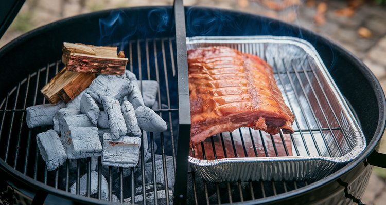 Two-zone charcoal grill setup with coals and wood on one side and meat above a water pan on the cool side