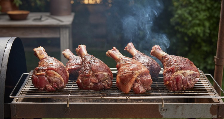 Turkey legs partially wrapped in aluminum foil on pellet grill grates with thin blue smoke