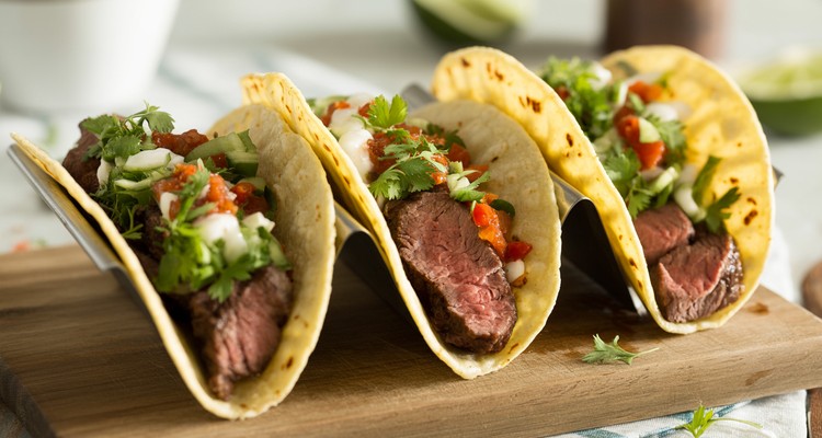 Three chuck steak tacos on a plate topped with diced onion, cilantro, and cotija with a lime wedge