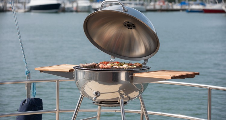 Magma stainless marine kettle grill mounted on a sailboat stern rail with seafood grilling over open water