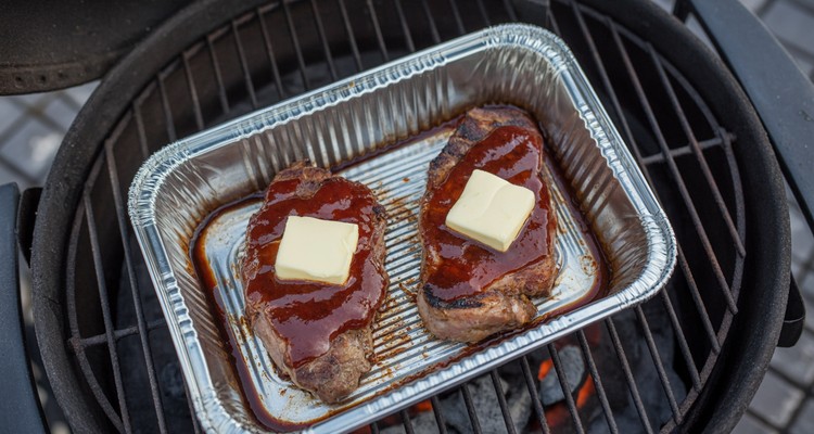 Aluminum foil pan on a smoker grate with smoked pork steaks topped with butter pats and BBQ sauce, ready to be covered with foil for braising