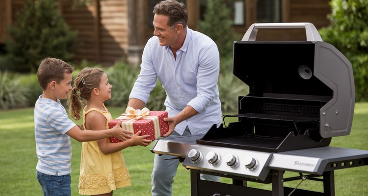 Wide hero view of a dad by a modern grill as a Father's Day gift in an outdoor setting with his kids giving him a wrapped present in the foreground, professional BBQ photography style, no text, no branding