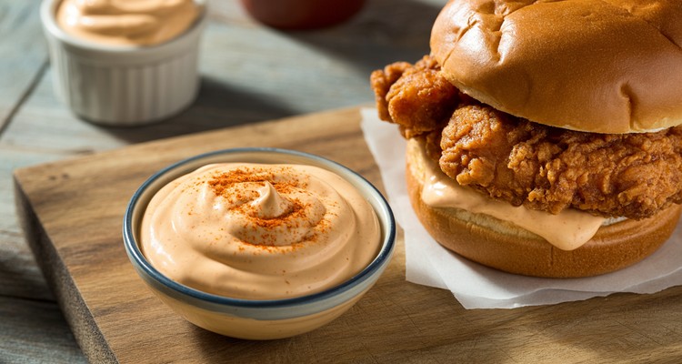A small bowl of creamy, orange-hued spicy mayonnaise garnished with a sprinkle of smoked paprika, next to a crispy fried chicken sandwich on a brioche bun