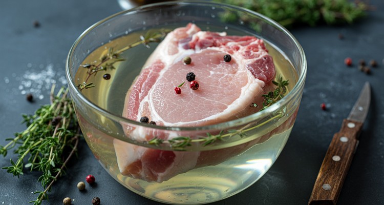Raw pork chops submerged in a clear glass bowl of brine with fresh thyme sprigs and black peppercorns floating in the water