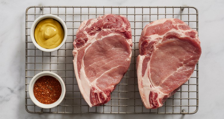Overhead shot of raw thick-cut pork steaks on a wire rack, one being brushed with yellow mustard and another coated in red rib rub